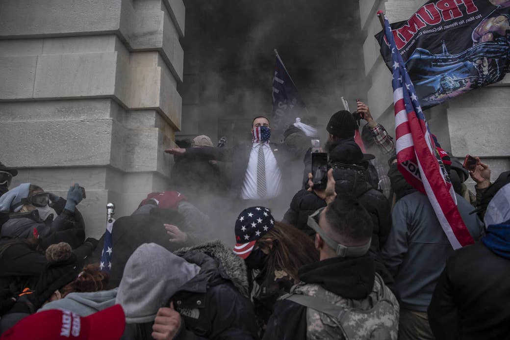 A man in a suit and a flag mask at the capitol riot with people and smoke all around him 