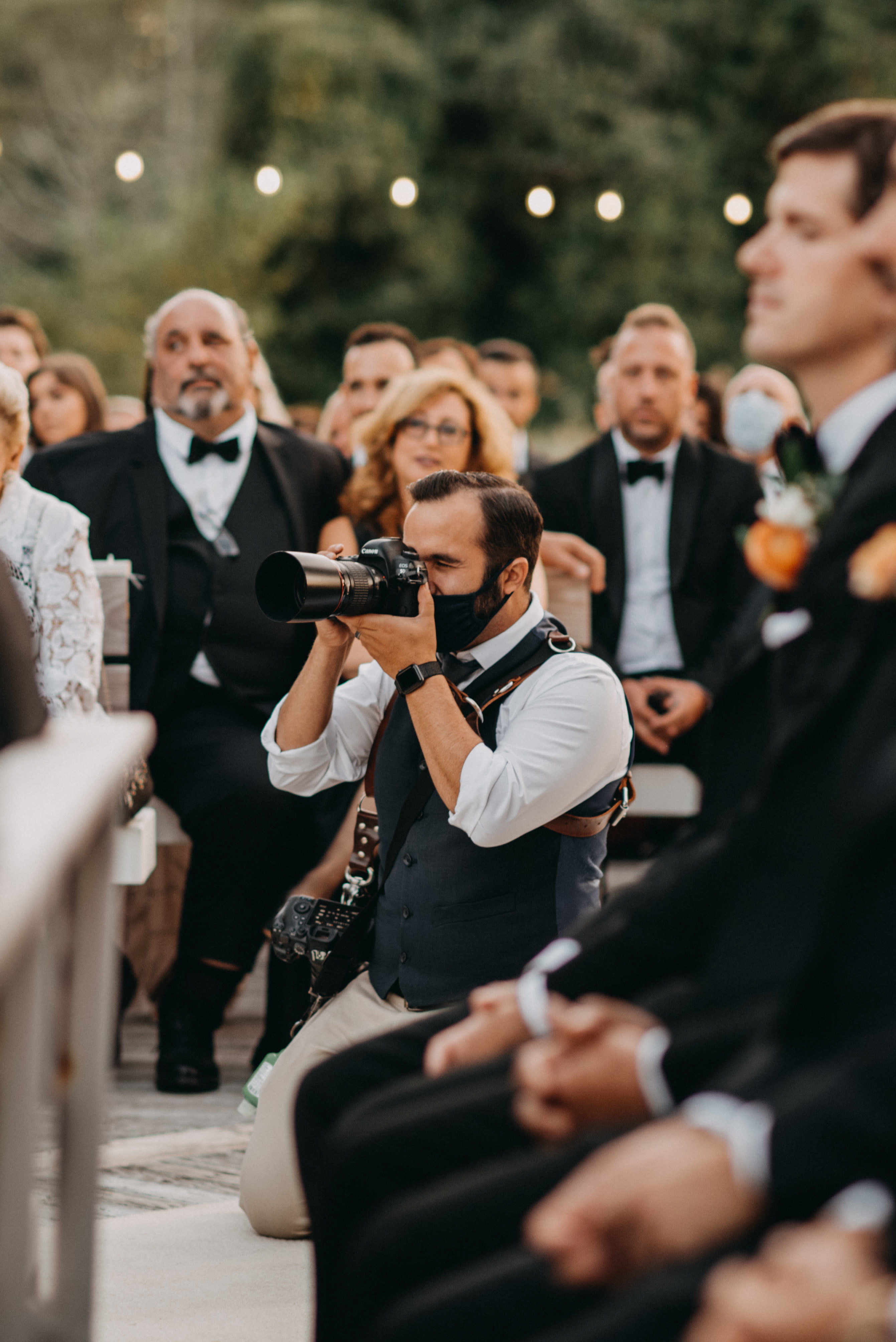 a wedding photographer with two cameras kneels down to take a picture surrounded by guests
