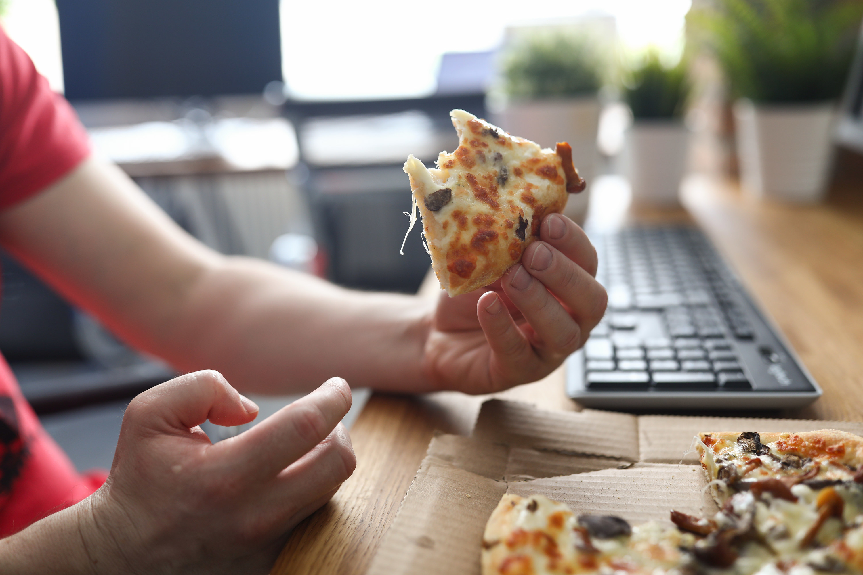 Hands holding a half-eaten slice of pizza at a desk with a keyboard