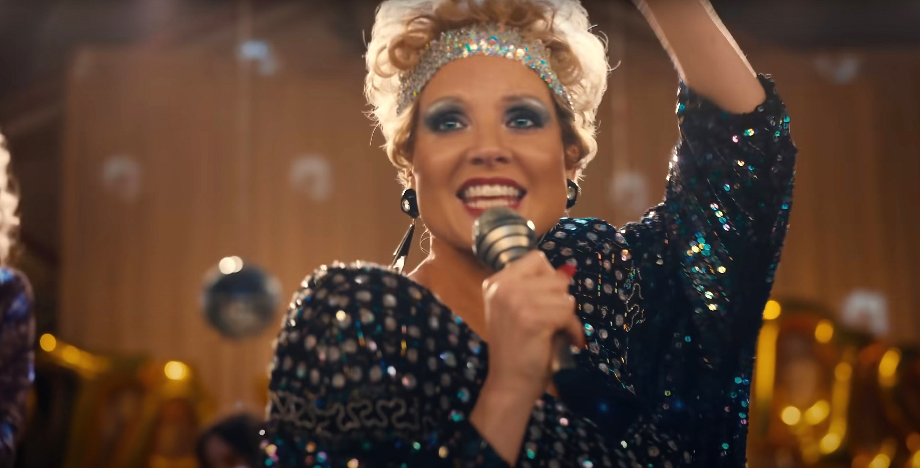Jessica sings into a microphone while playing Tammy who is wearing a sequin headband around short curly hair