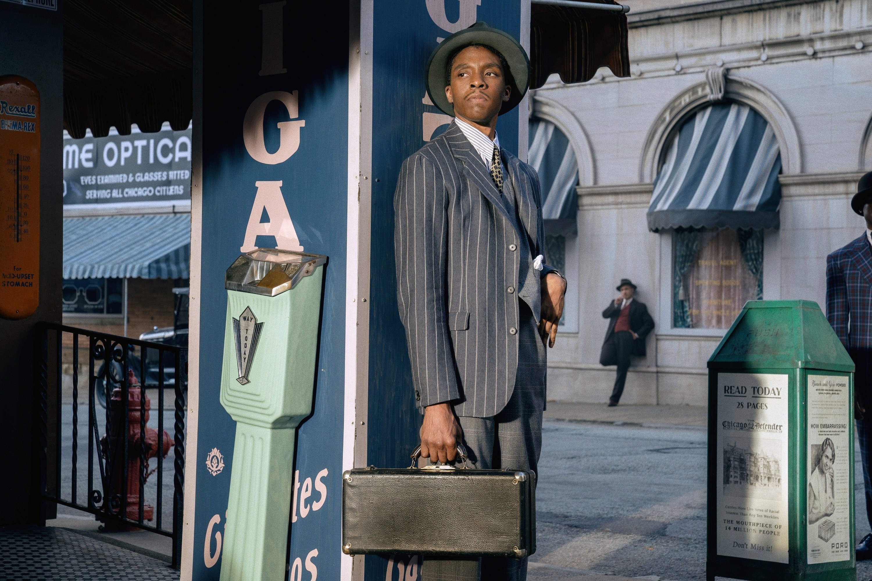 Boseman stands next to a parking meter while wearing a suit and carrying a suitcase