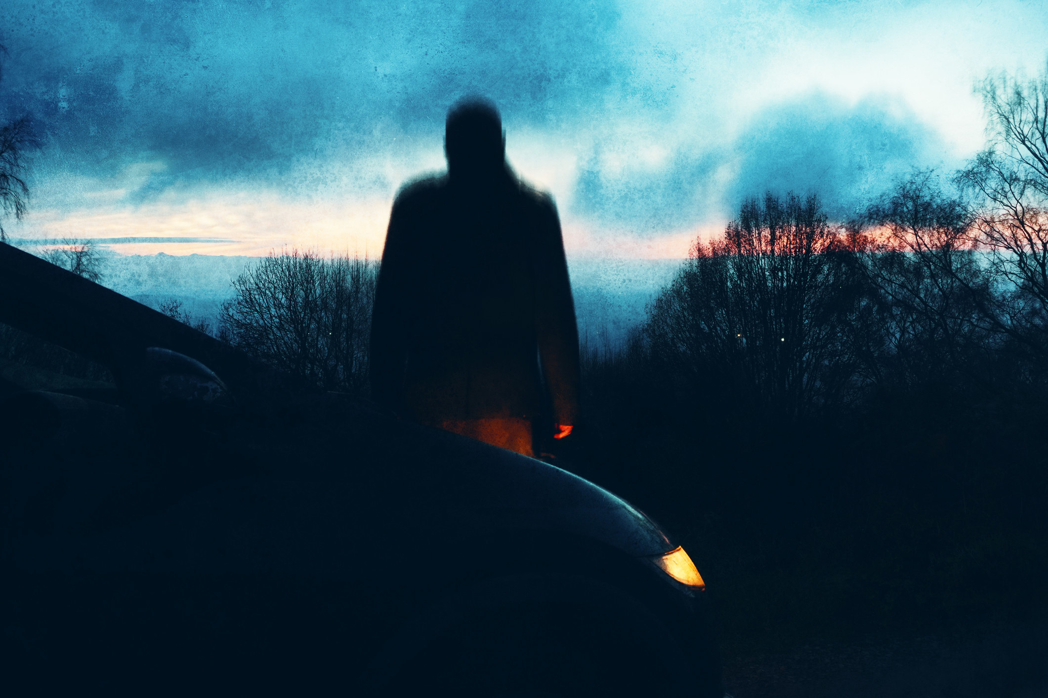 A silhouette of man standing next to a car during the early hours of the day