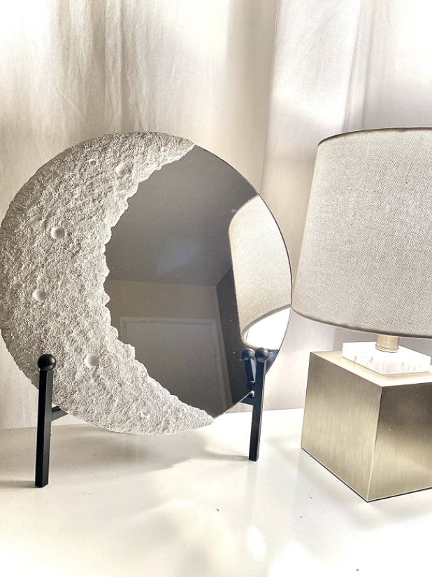 round mirror with realistic looking crescent moon design on one side. it is balanced on a stand and placed on a desk.