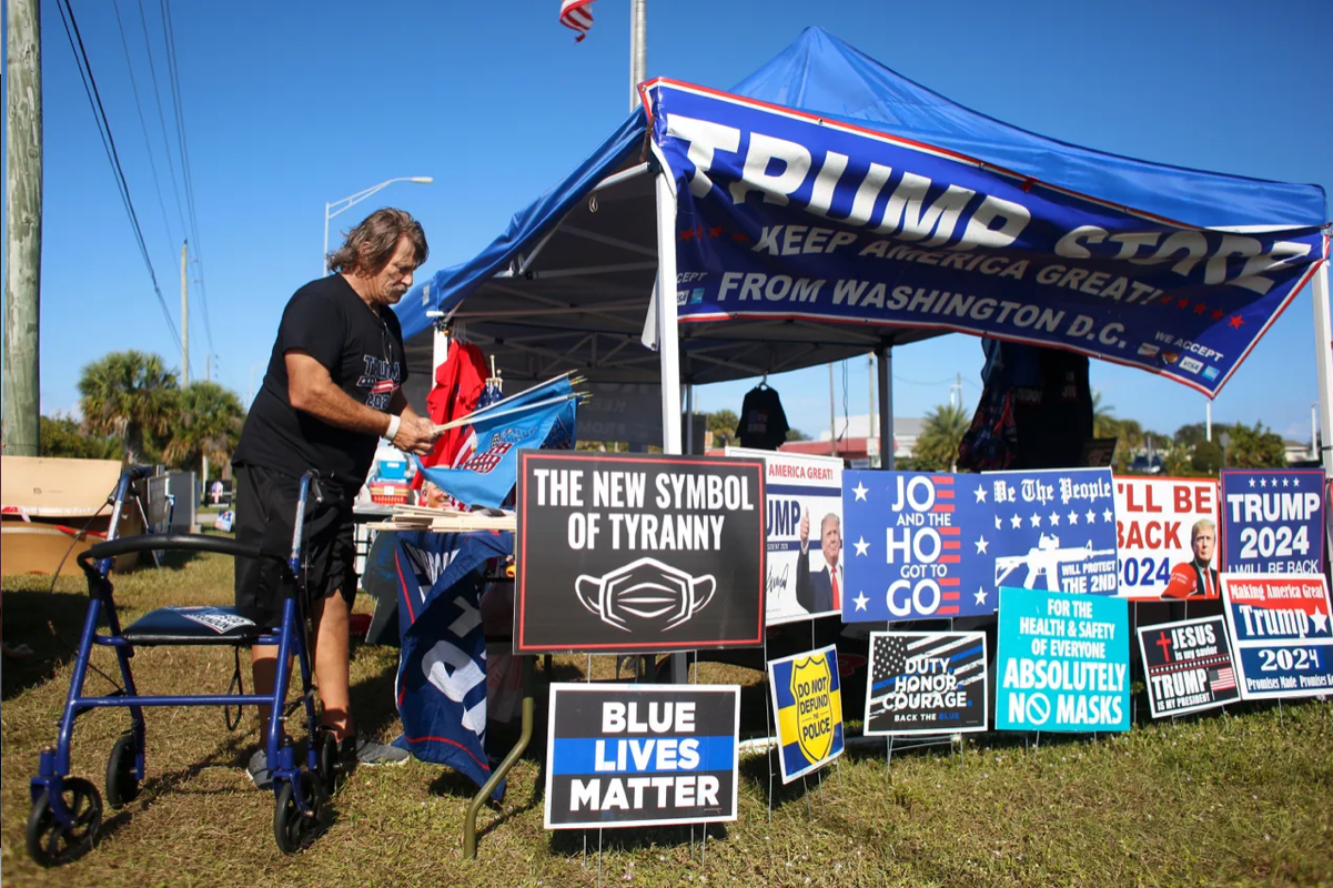 A man with a walker and a bunch of signs for trump and blue lives matter near a tent 