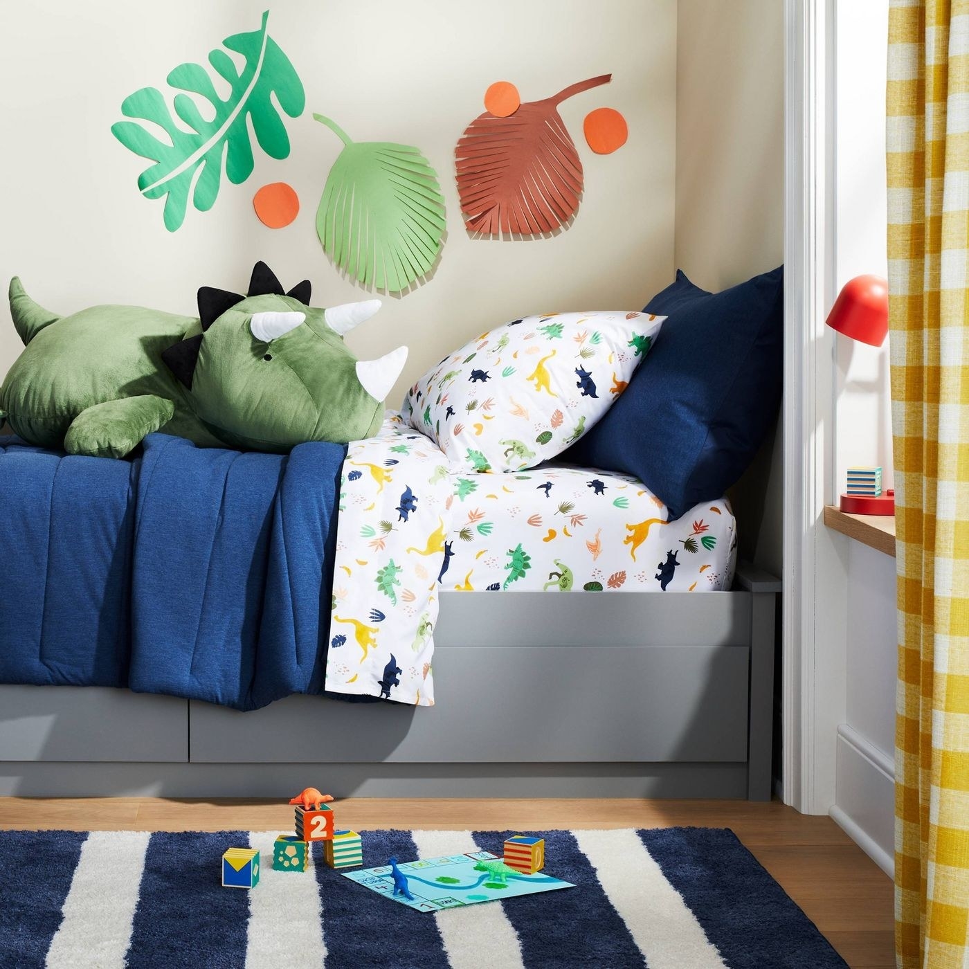 A kid&#x27;s bed with white sheets that have a dinosaur print, along with a navy comforter and green dinosaur stuffed animal.