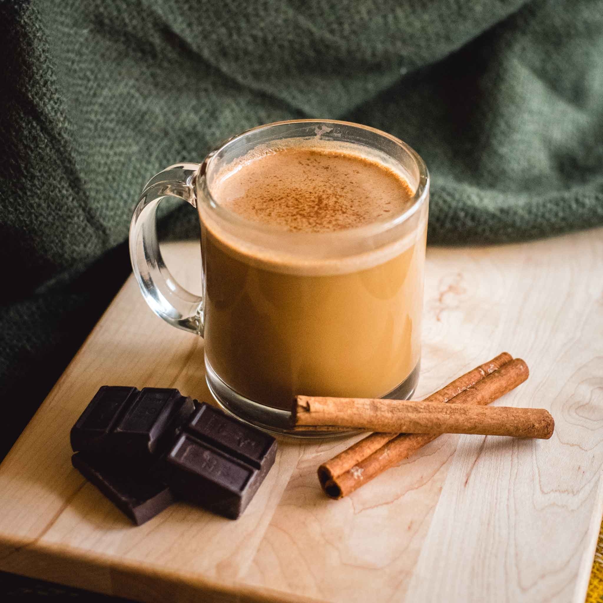 A cup of the mushroom hot chocolate next to chunks of chocolate and cinnamon sticks