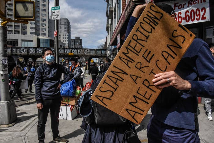 A person wearing a mask stands in the background as a person holds up a sign saying &quot;Asian Americans are Americans!&quot;