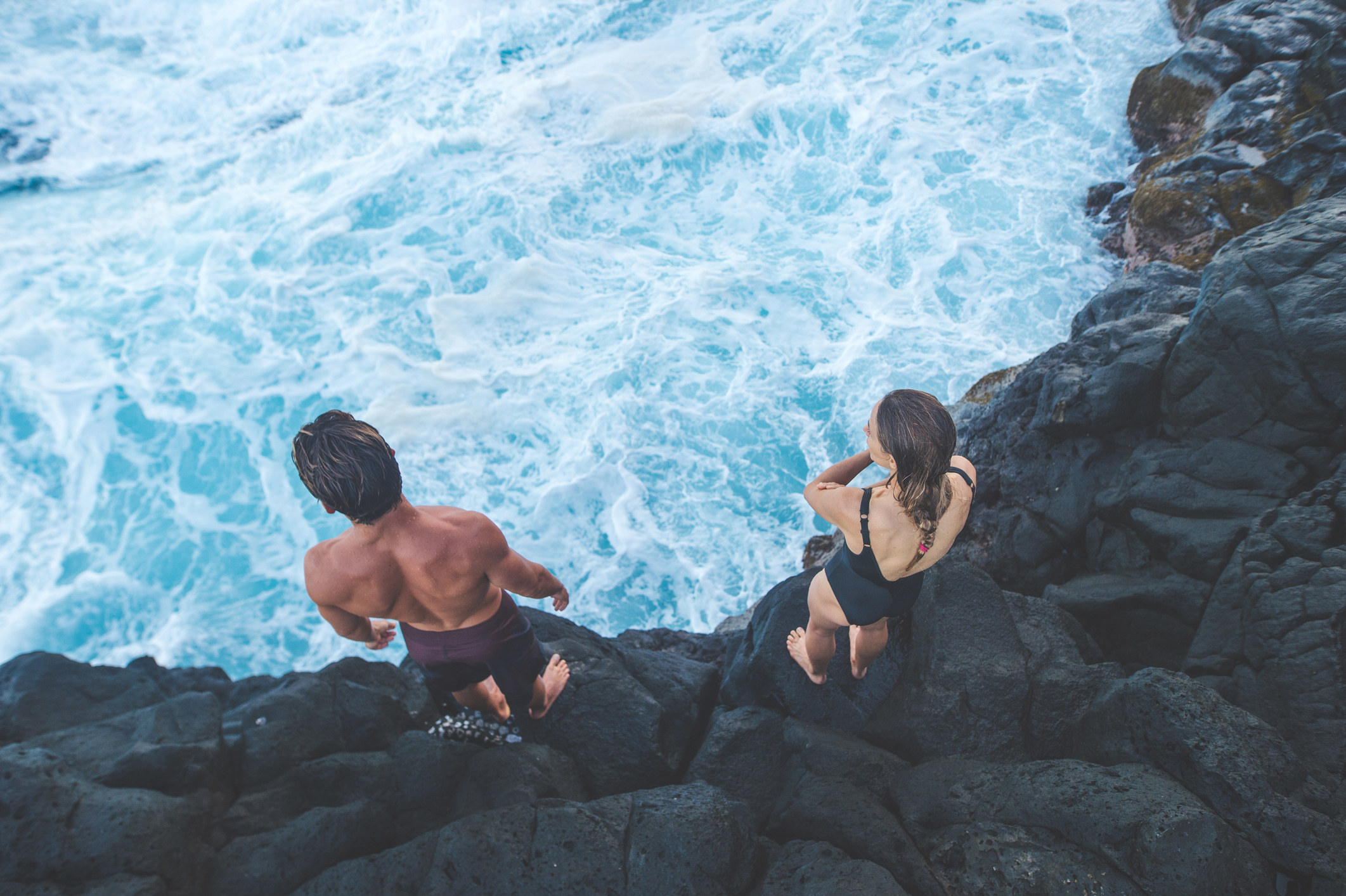Two people in bathing suits standing on a cliff looking at the water below
