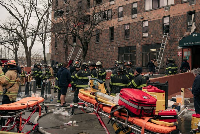 Many firefighters and emergency responders stand outside a brick building, with several ladders leaning against it