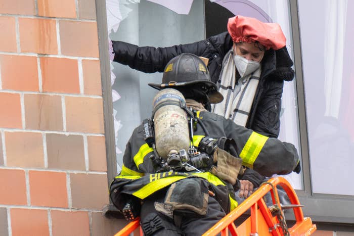 A firefighter stands on a ladder up against a broken window, while a person emerges from it