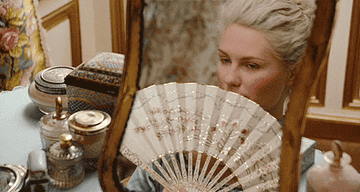 Marie looking into a mirror on her vanity as she holds up an ornately decorated fan