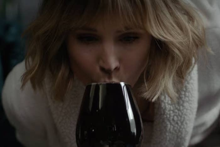 Anna is bent over a counter while sipping wine from a glass.