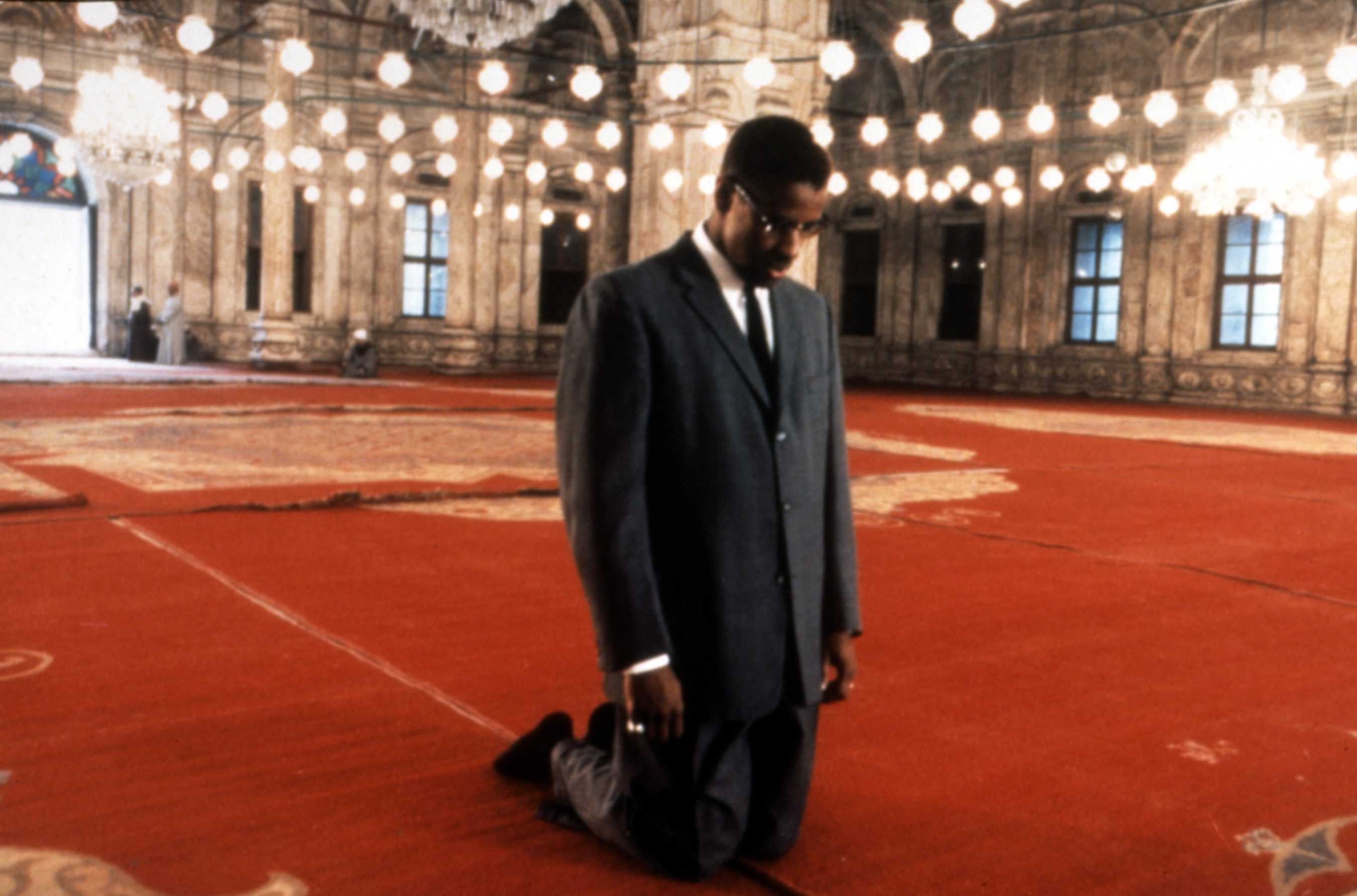Malcolm X kneels and bows his head inside a house of worship