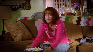 Frankie Heck from &quot;The Middle: sitting on a brown sofa, in grey tracksuit bottoms and a pink hoodie. She grabs a bowl of popcorn and then starts to eat it while watching something excitedly.