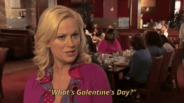 Woman with short blonde hair, wearing a pink blouse and pink cardigan standing in a restaurant. She&#x27;s saying &quot;What&#x27;s Galentine&#x27;s Day? Oh, only the best day of the year&quot;