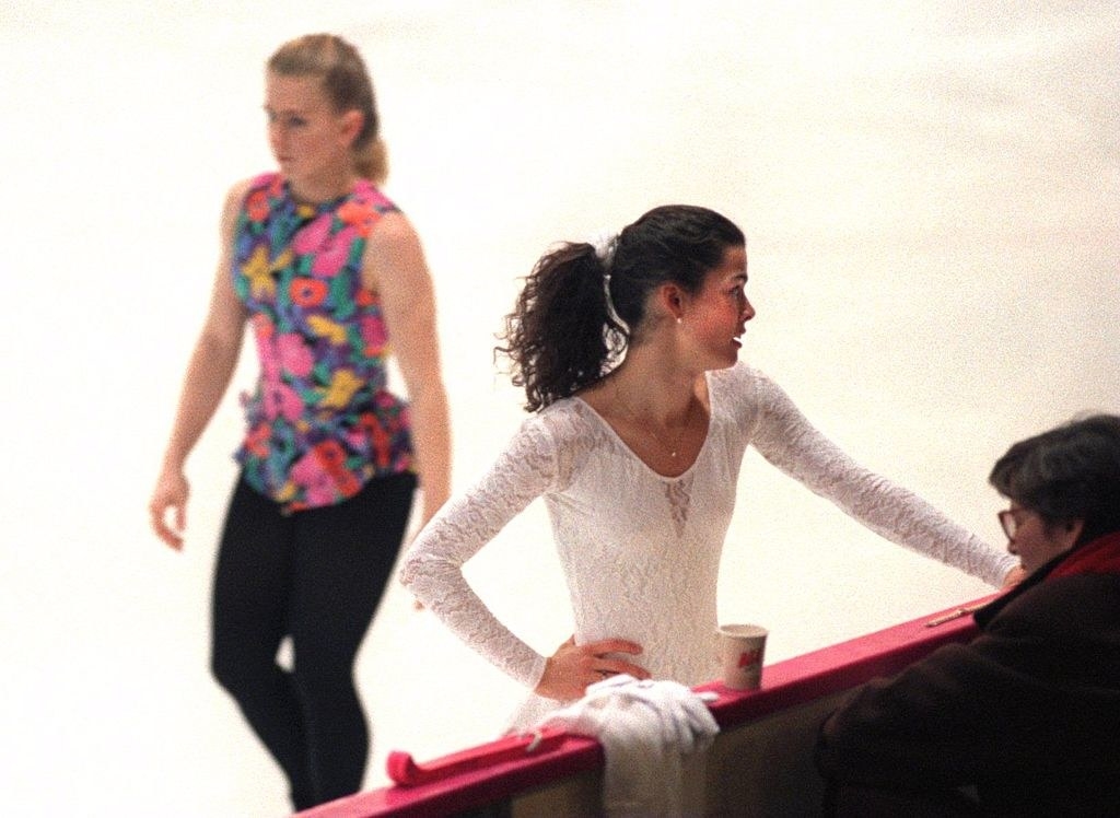 Nancy stands at the edge of the skating rink as Tonya Harding skates by in the background
