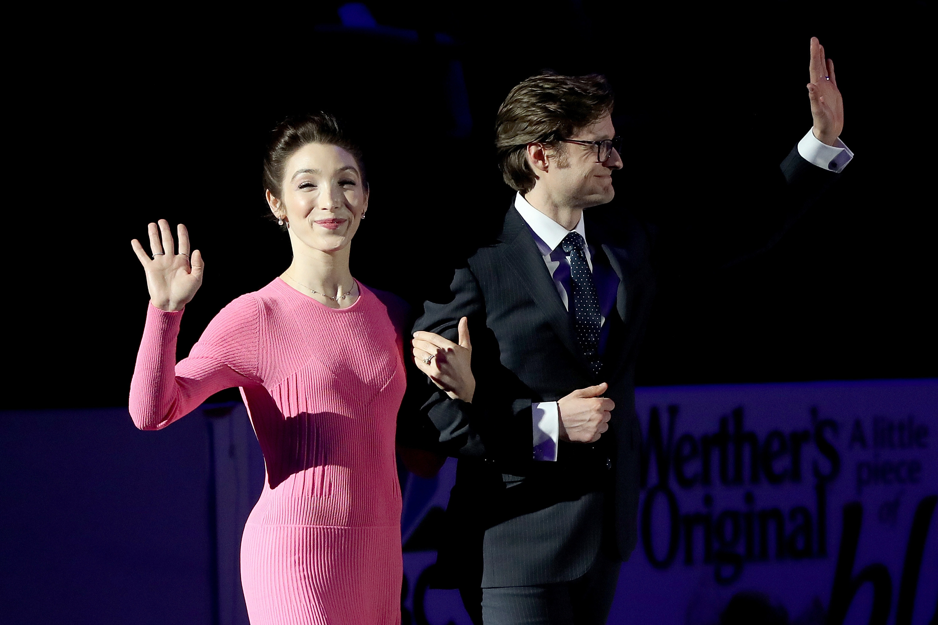 the two, in a suit and dress, waving to a crowd while on ice