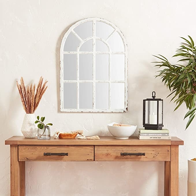 The mirror hanging on a wall above a console table with plants, candles, and knickknacks on it and a palm plant beside it