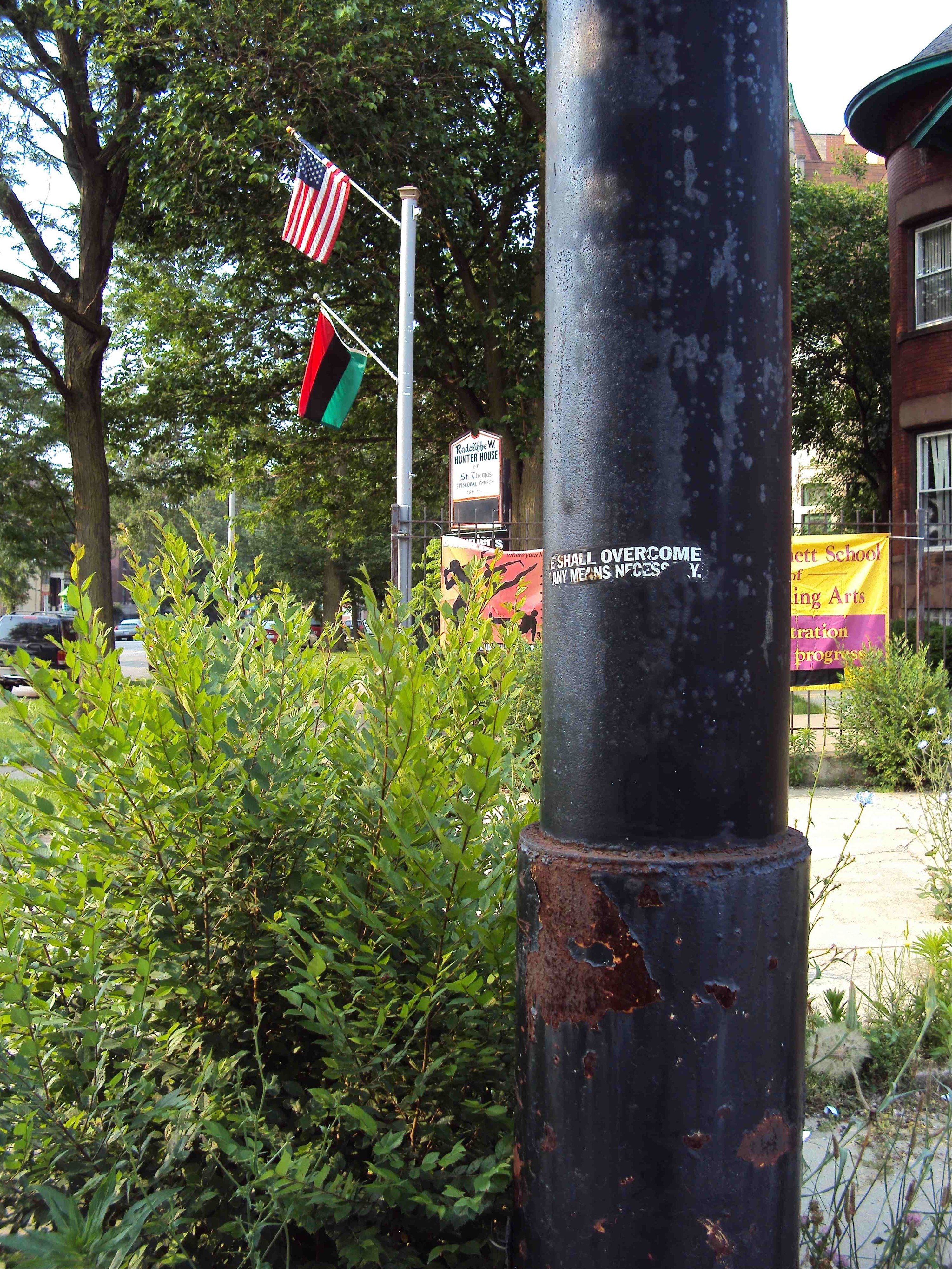 a photograph of a lampost with a torn bumper sticker that says we shall overcome and flags in the background