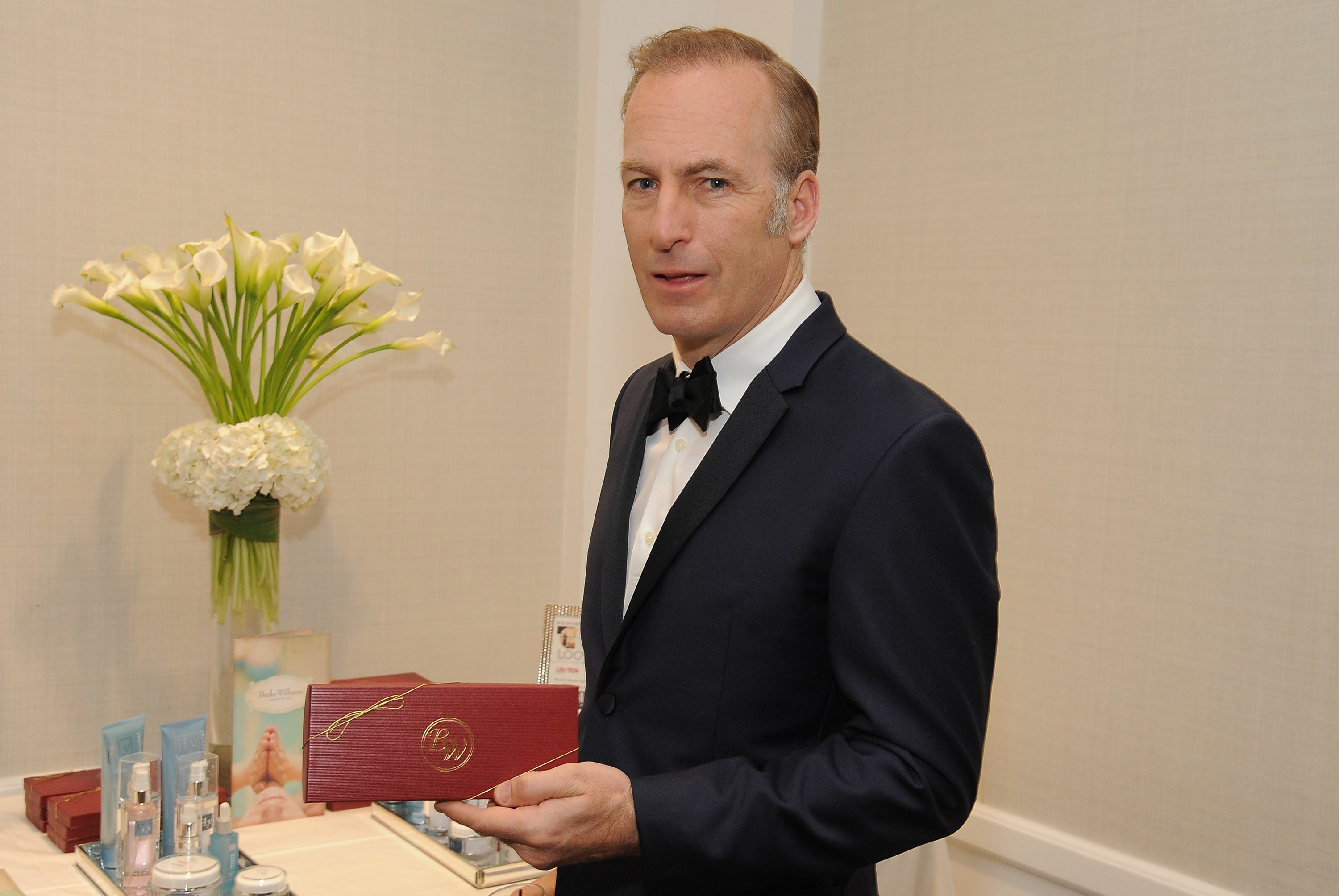 Odenkirk holds a red box while standing in front of products