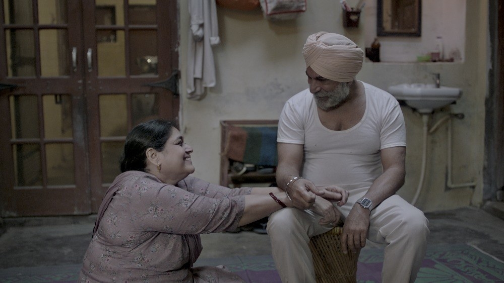 Supriya Pathak and Pavan Malhotra smile at each other. Supriya is sitting on the floor while Pawan sits on a wooden stool