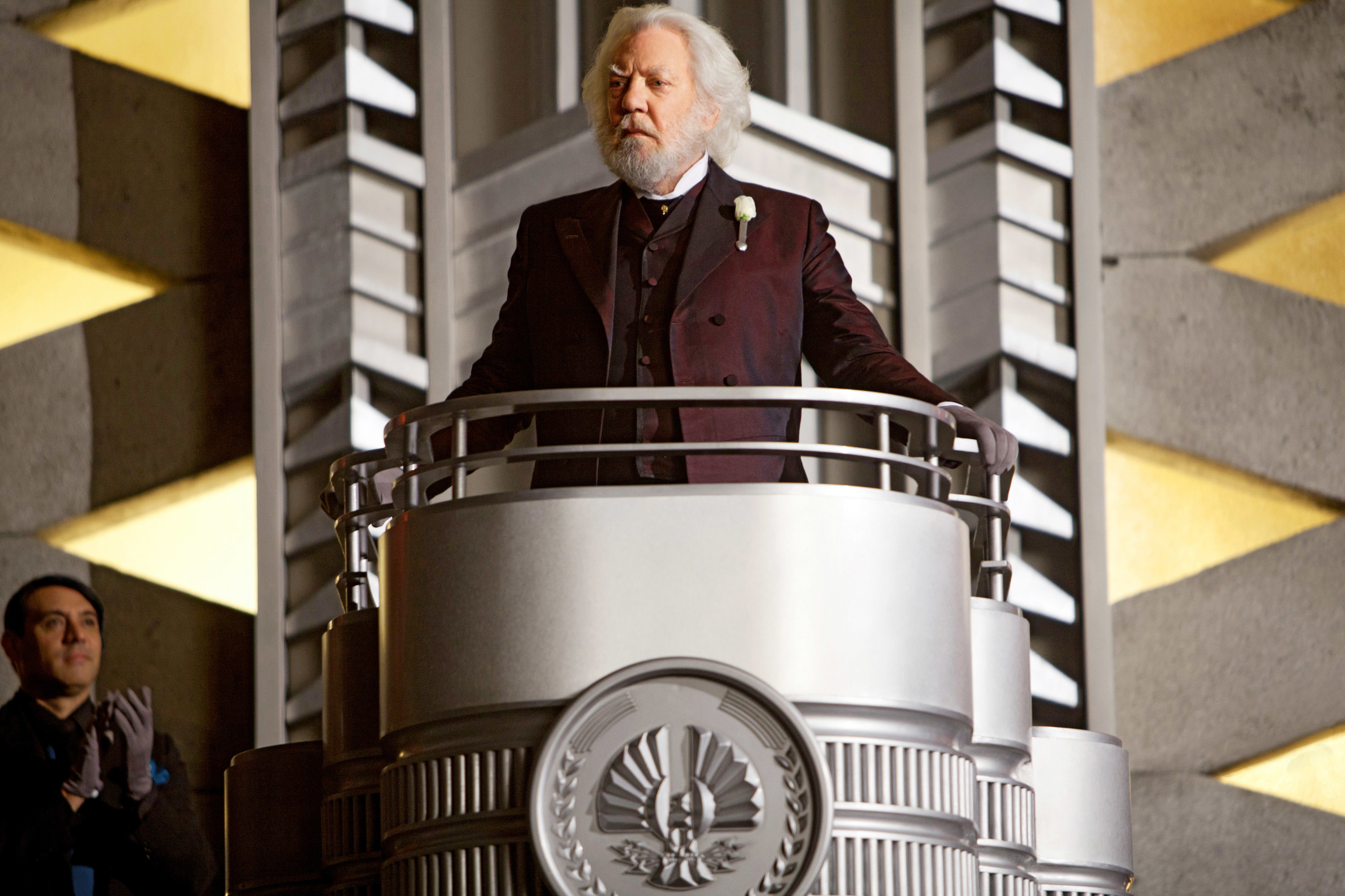 President Snow in a mottled red and black suit with a white rose pinned to his lapel