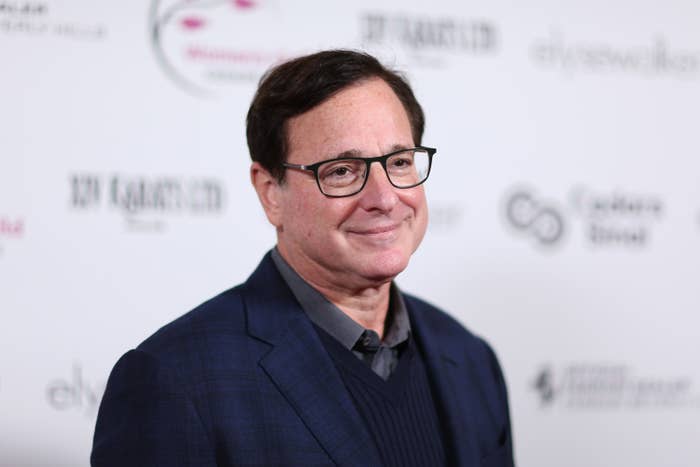 Bob Saget from the shoulders up in a suit and v neck in front of a background with indistinguishable writing
