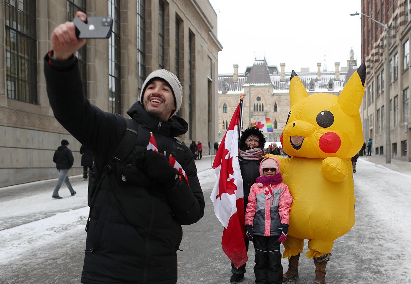 A man stands in the street and takes a selfie with a woman holding a Canadian flag and a child smiling behind him