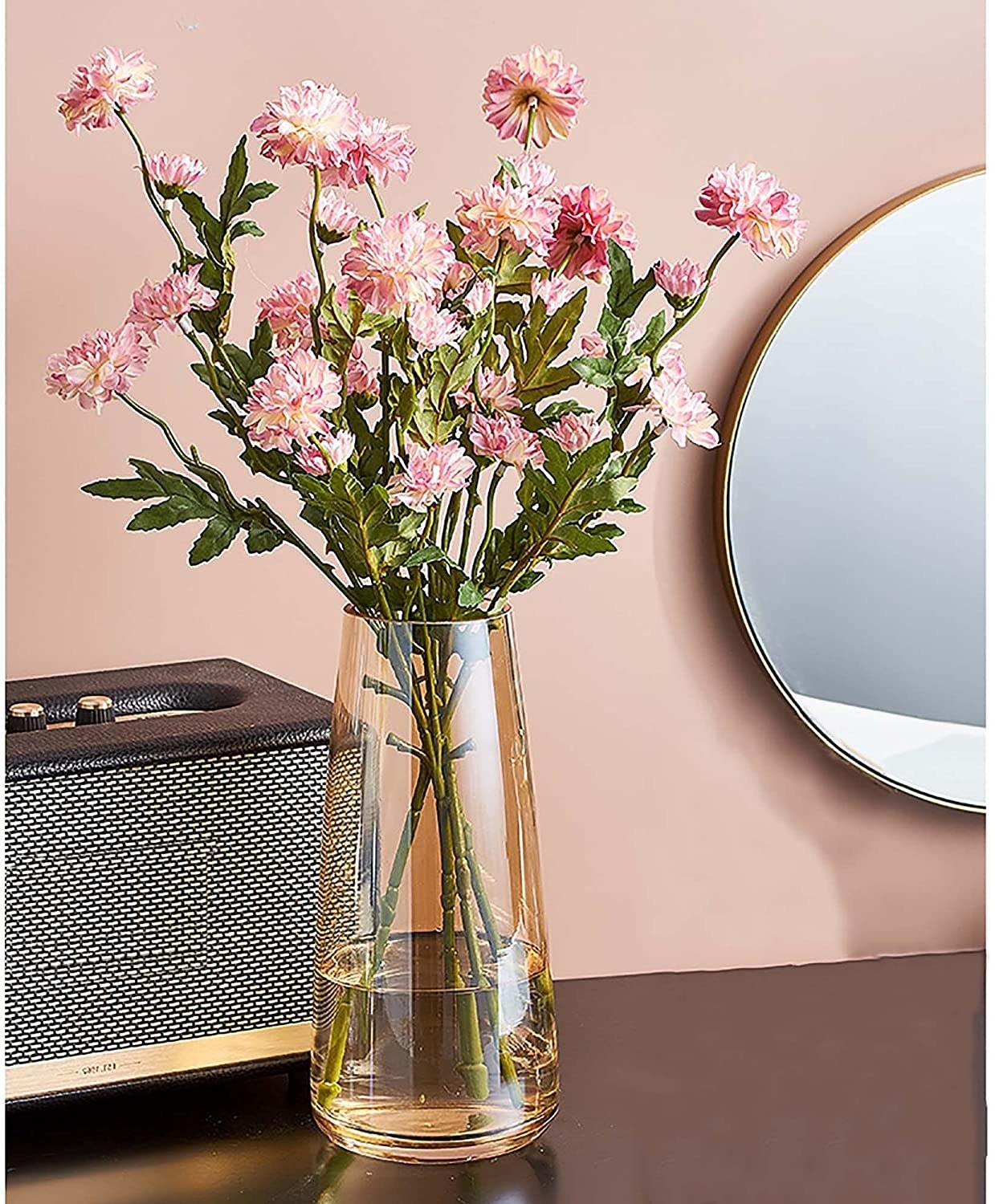 A glass vase filled with flowers sitting on a table in front of a small speaker and mirror