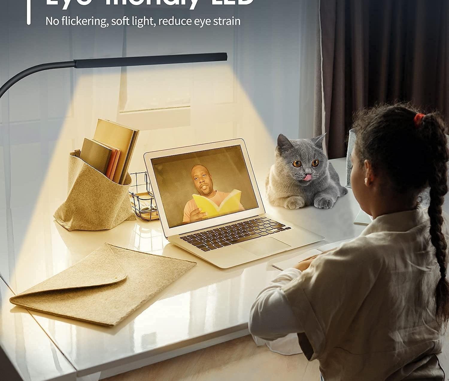 A child sits at a desk with a laptop, school supplies, and a cat sitting on it while they are on a video call with an adult