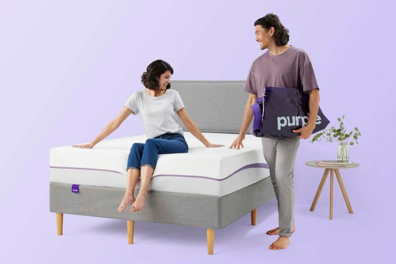 a woman sitting on a purple mattress with a man standing next to it with other purple products