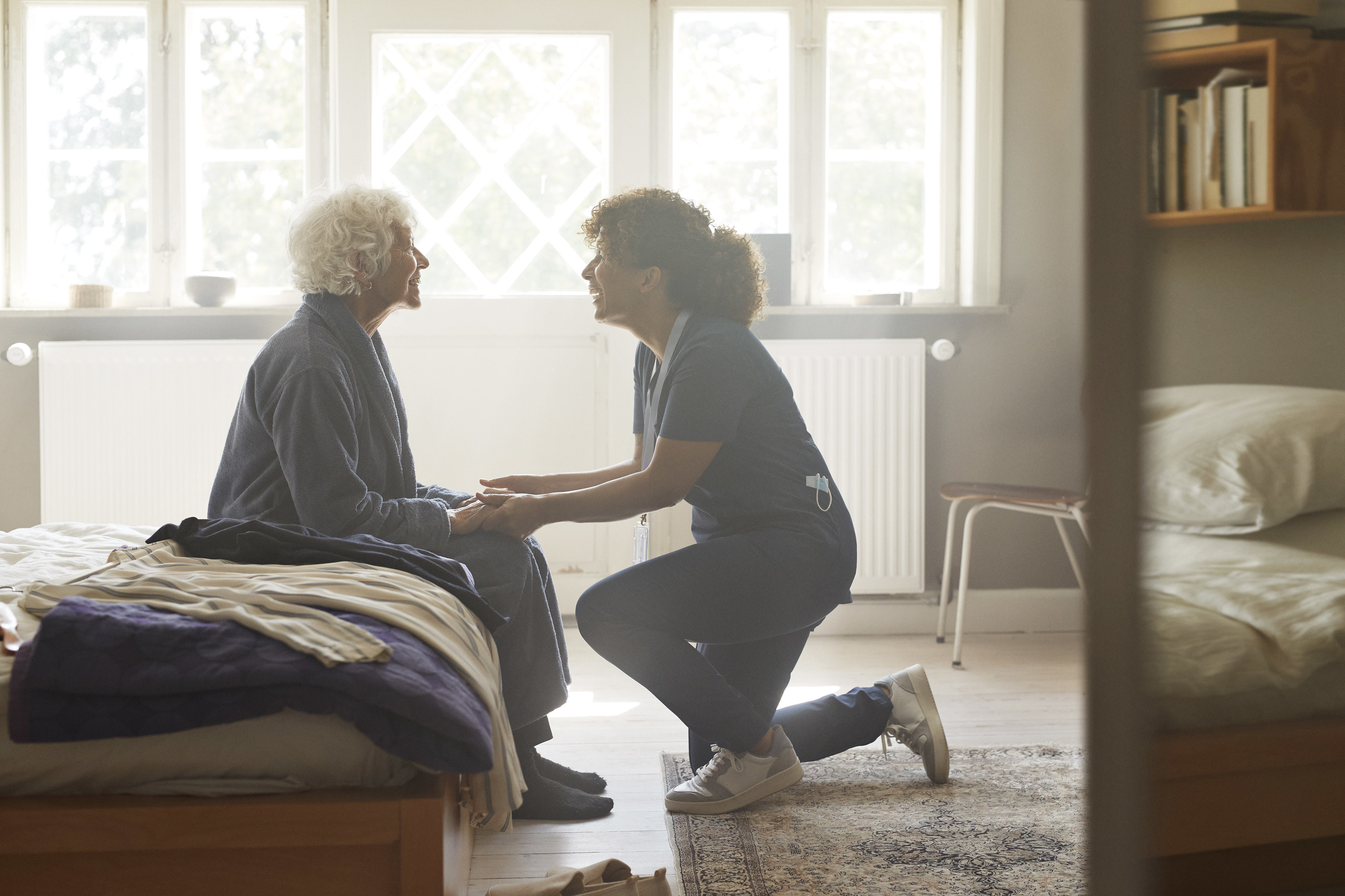 a nurse kneeling as she holds the hands of an old woman sitting on the edge of her bed