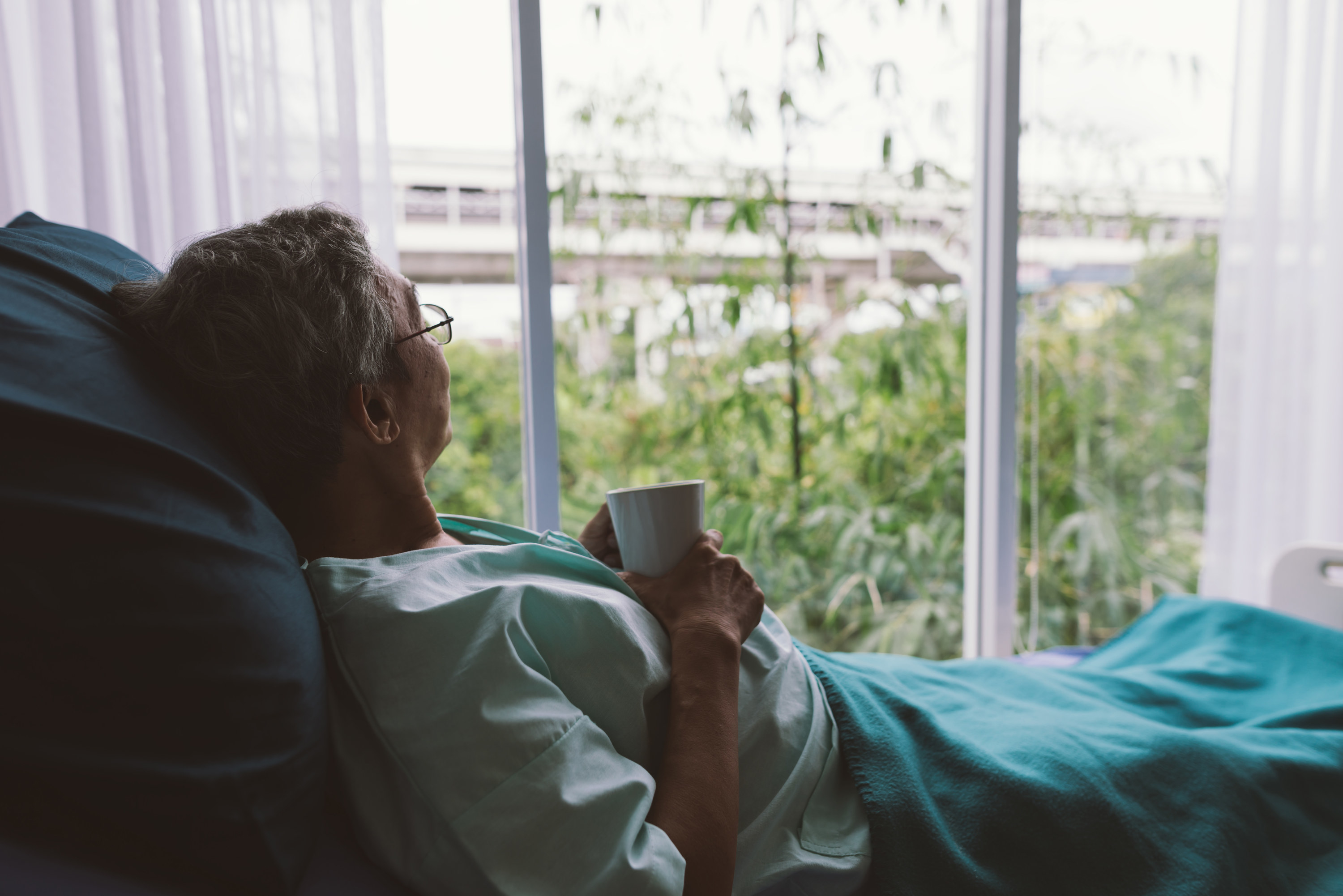 an elderly man drinks tea while looking out the window from his hospital bed
