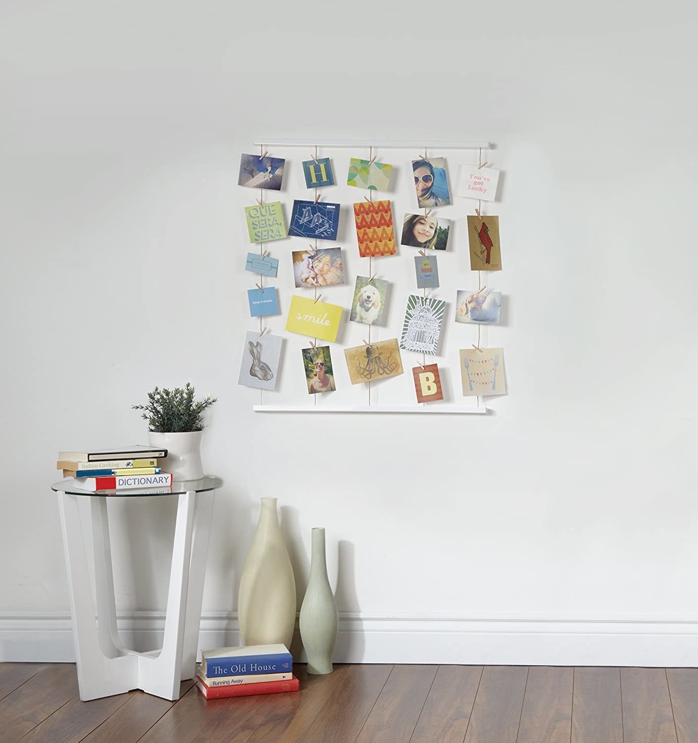 The hanging collage kit hanging vertically on a plain wall next to a small table with books on it, and vases on the floor