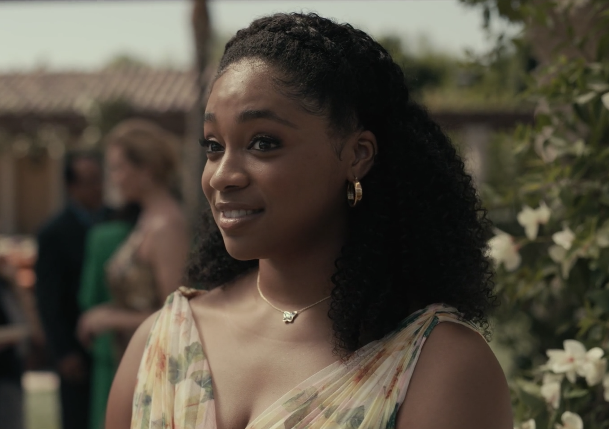 Simone Joy Jones as Lisa, wearing a white dress with a floral pattern, a butterfly necklace and gold hoop earrings