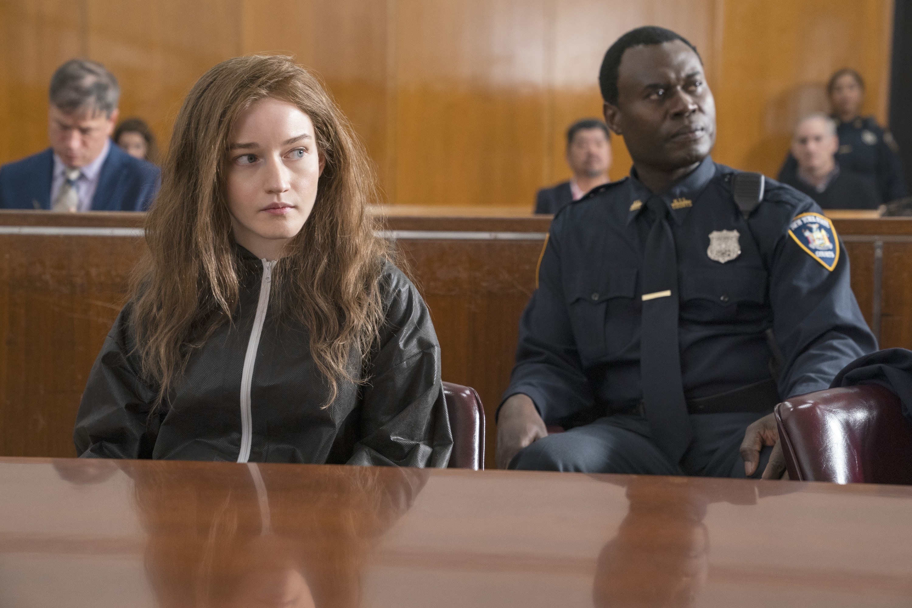 dressed in a zipped-up jacket with messy hair, Anna sits next to a guard in court