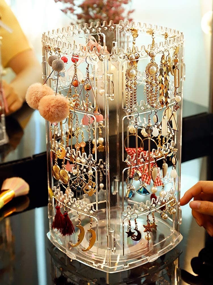The four-sided earring stand on a table holding lots of earrings an a person&#x27;s hand on it about to turn it