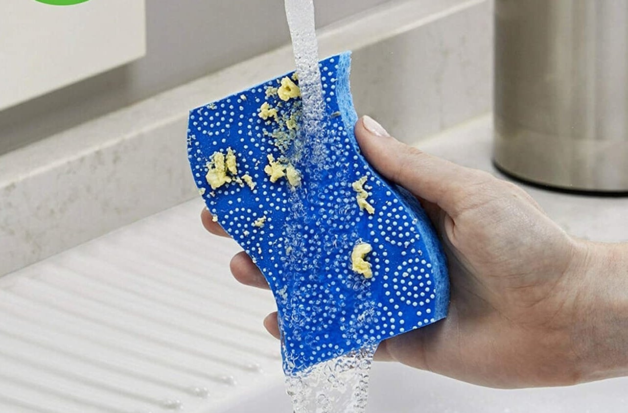 A person holding the sponge under a running faucet with food particles stuck to the little grippy dots on the sponge