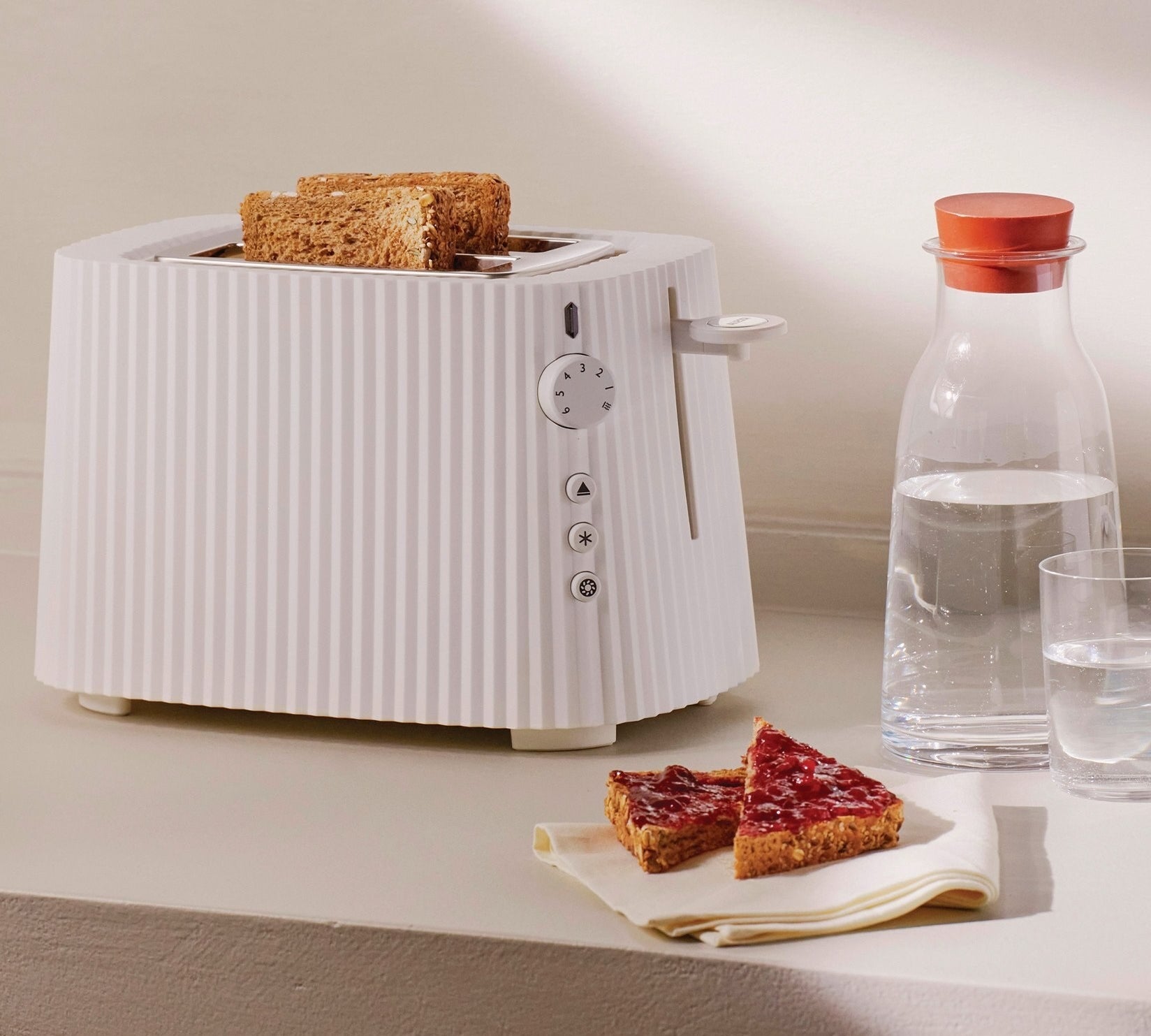 the white pleated toaster holding two pieces of toast and placed next to some jam-covered pieces of toast and water