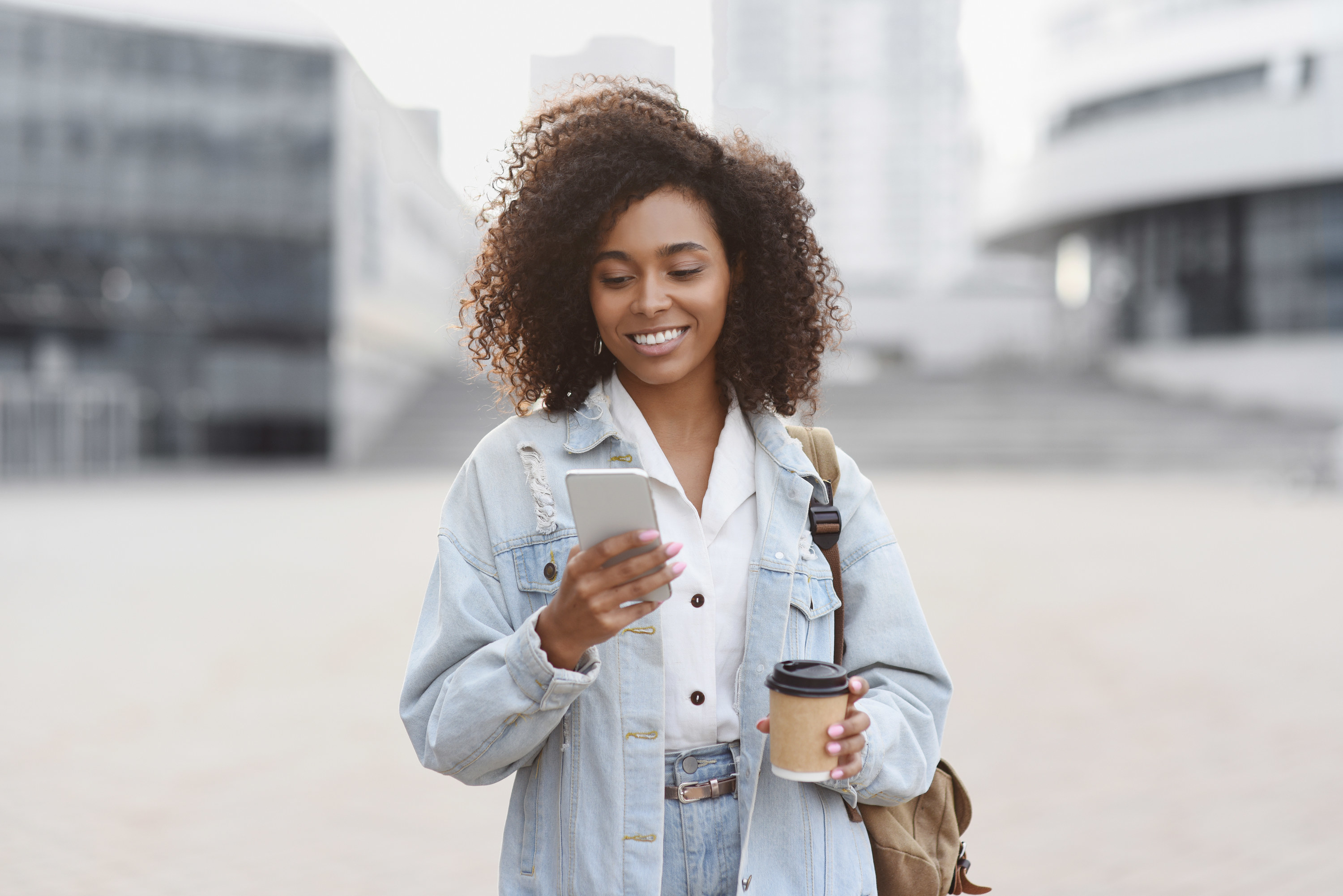 A woman staring at her phone with a smile on her face while carrying a coffee
