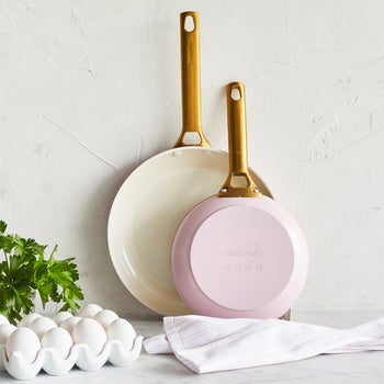 the two pink pans standing up against the wall on top of a kitchen counter, next to a towel, eggs, and herbs