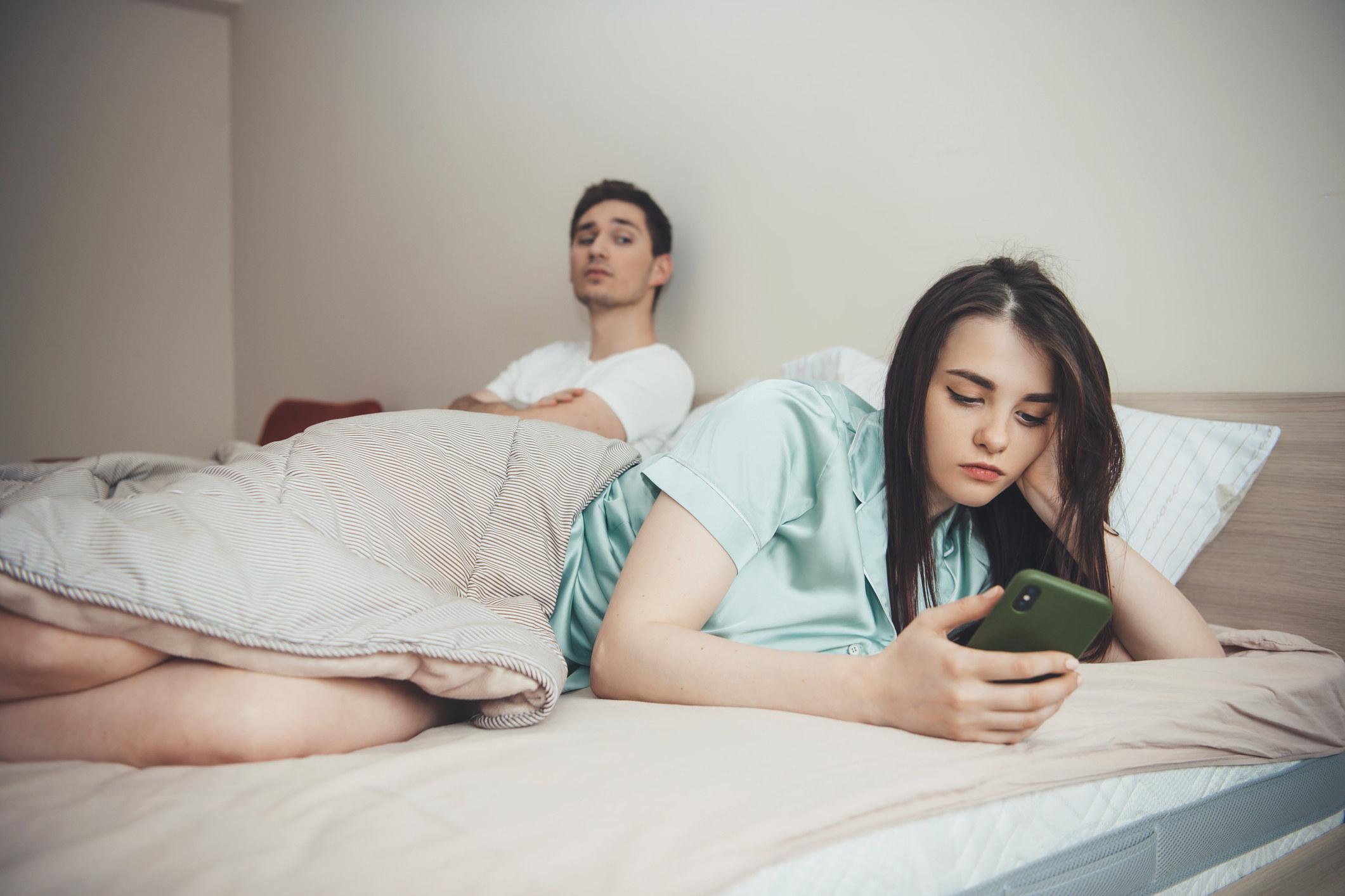 Jealous man watches over his girlfriend&#x27;s shoulder as she texts in bed