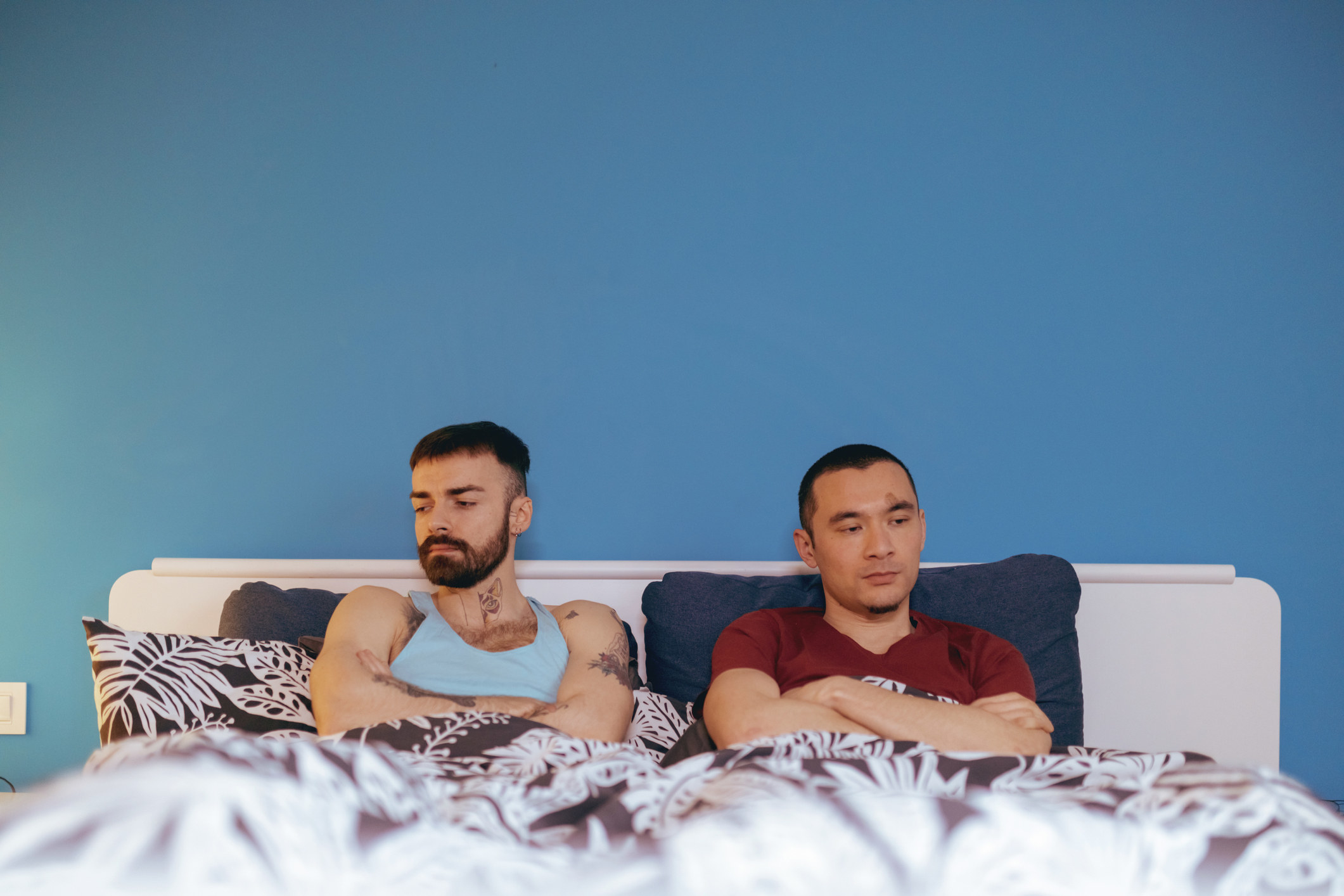 Displeased gay couple sitting in bed under the sheets with arms crossed after an argument