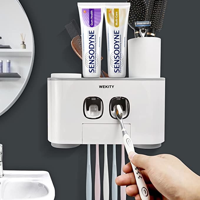 The teeth cleaning station hanging up on a bathroom wall while a person reaches a toothbrush into it to dispense toothpaste onto the brush