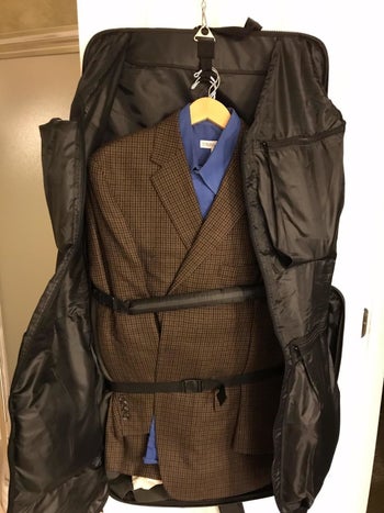 same reviewer's garment bag hanging up showing the inside with pockets and three outfits on hangers strapped inside