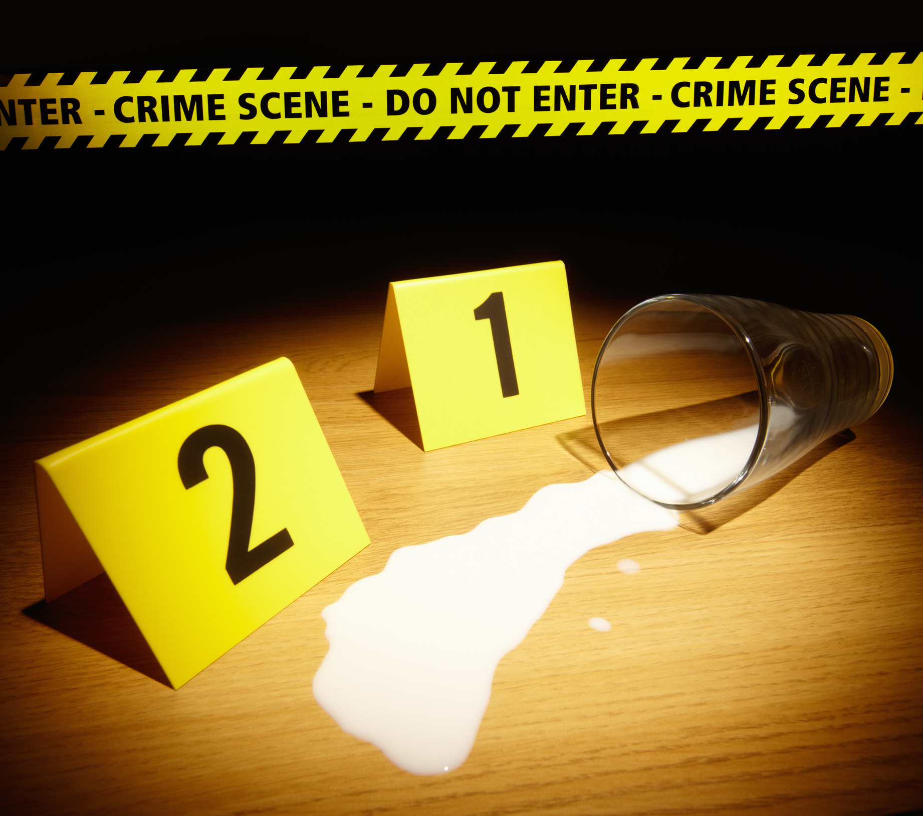 Glass of spilt milk with crime scene tape and crime scene numbers on tabletop