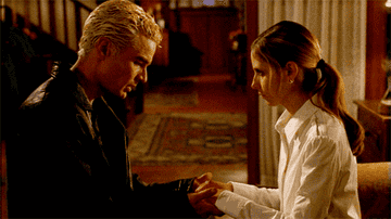 Buffy and Spike hold hands as they sit in a living room