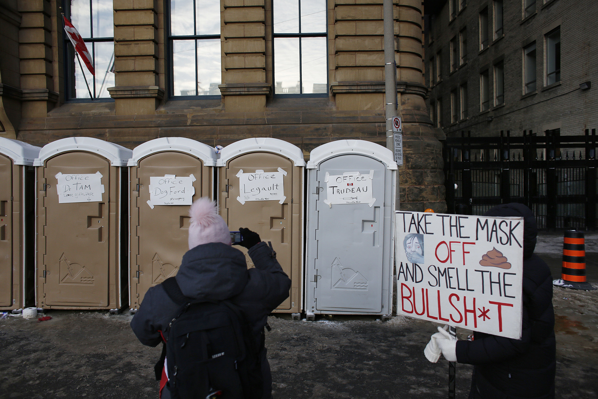 A person standing by a sign reading &quot;take the mask off and smell the bullshit&quot; takes a cellphone image of porta-potties, each with a taped-up sign denoting it as the office of a Canadian politician