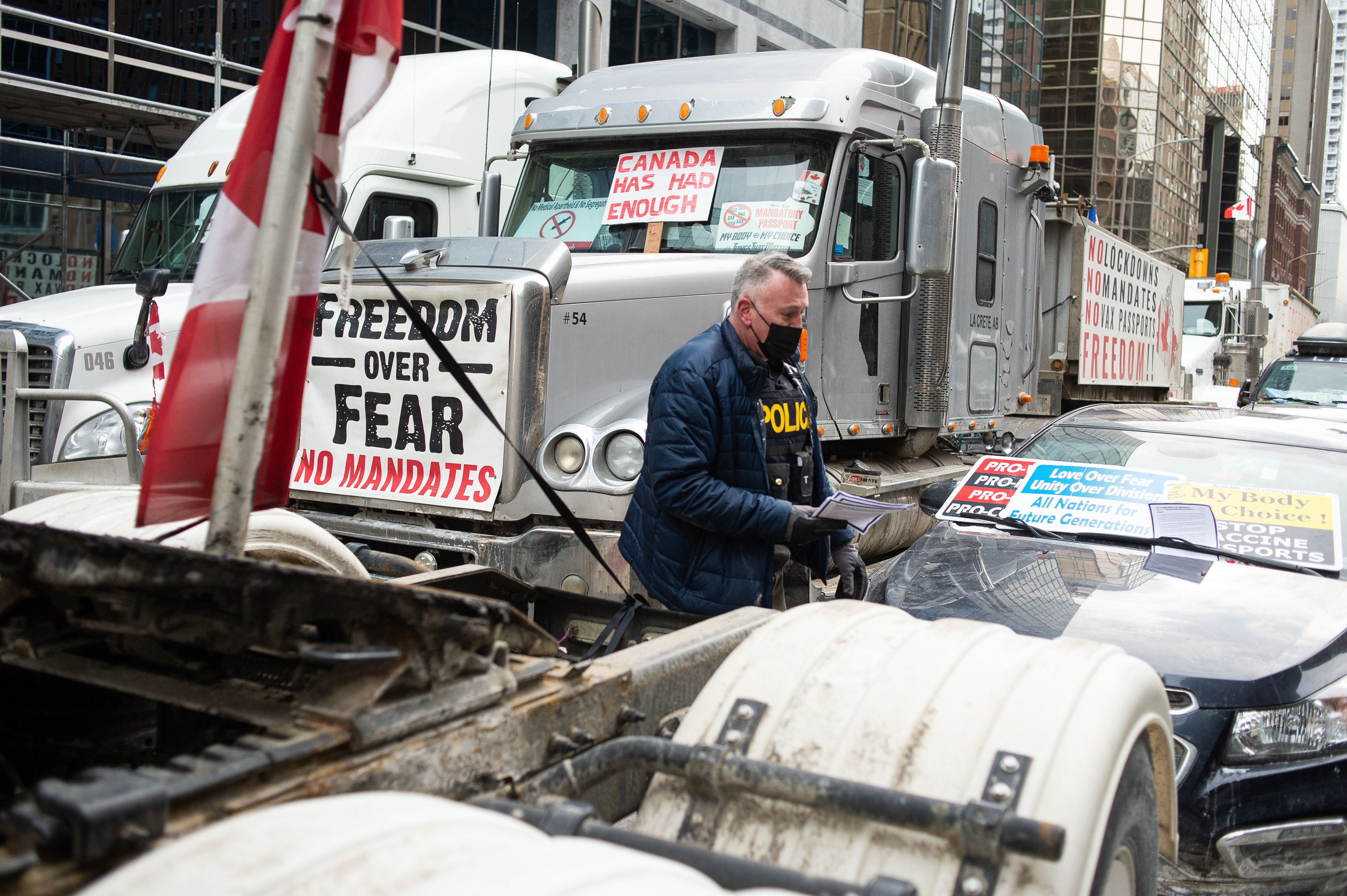 An officer wearing a vest labeled &quot;police&quot; carries a stack of flyers and places on under a windshield beside a big rig with signs posted reading &quot;freedom over fear, no mandates&quot; and &quot;Canada has had enough&quot;