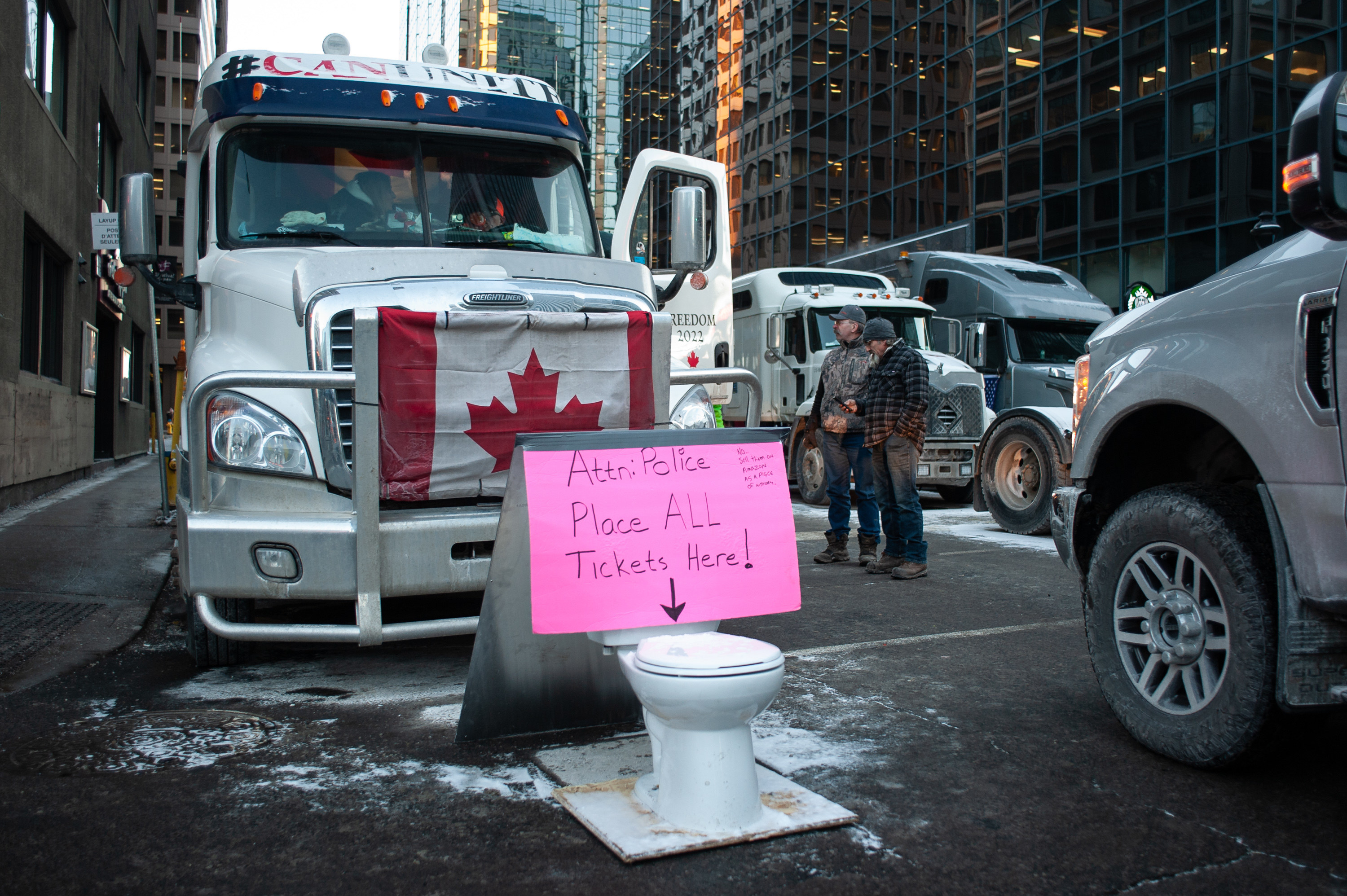 Cars and big rig trucks are parked in the street, where a toilet sits in the road with a sign reading &quot;attention police, place all tickets here&quot; with an arrow pointing into the toilet bowl